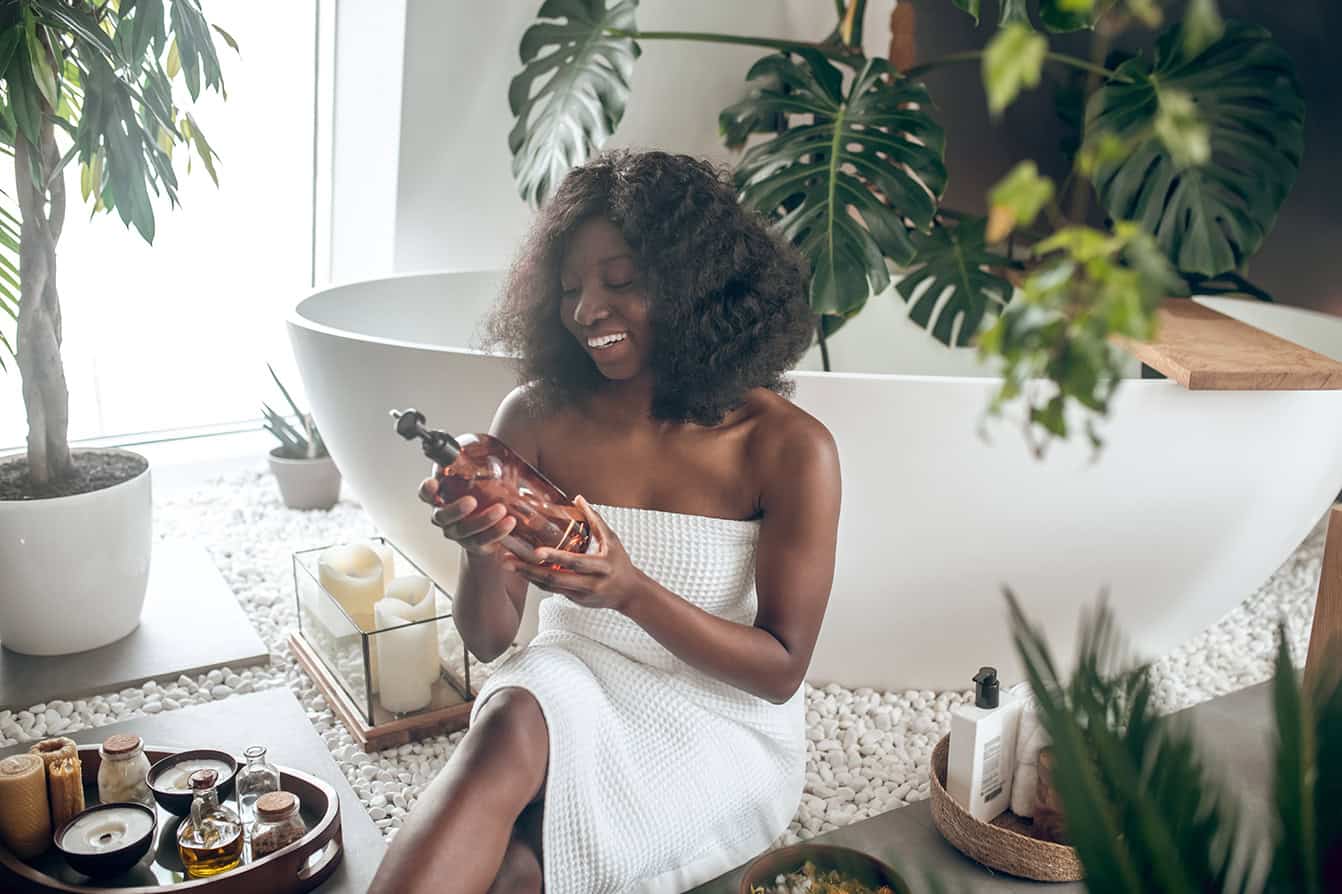 Woman in towel holding bottle beside bathtub