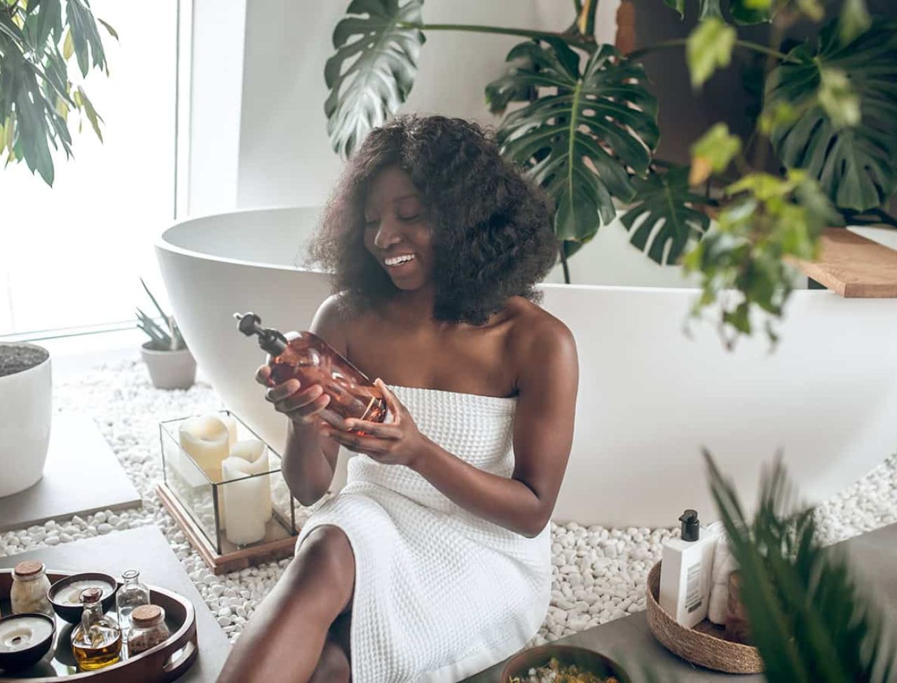 Woman in towel holding bottle beside bathtub
